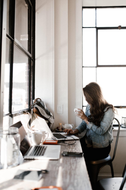 A young woman sitting at a desk near a bright window,
                    working on her laptop and drinking a cup of coffee.